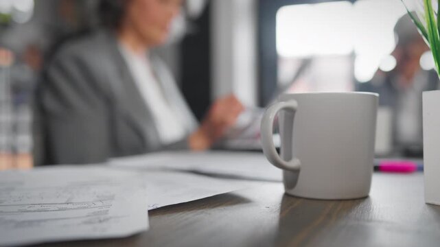 Cozy workplace in office, aged woman checking reports and accounting profit. Ceramic cup on table in foreground, multitasking senior lady in background, working day in office, professional accountant