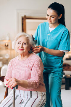 Smiling positive caregiver combing senior woman's hair in home