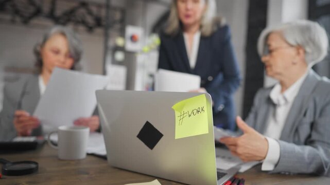 Modern co-working space, working day in office, senior woman working with laptop . Yellow sticky note on computer with work inscription, elderly female person using pc for job, career for ladies