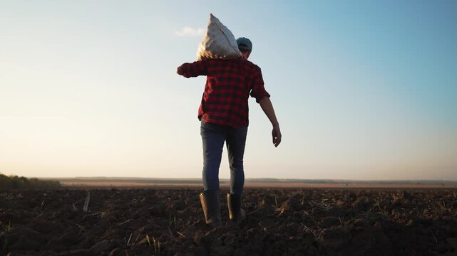 Farmer carries heavy sack across plowed field. Worker walks on soil at sunset. Farmer in boots steps over dirt. Plowed field stretches to horizon. Agriculture worker hauls seed bag on shoulder.