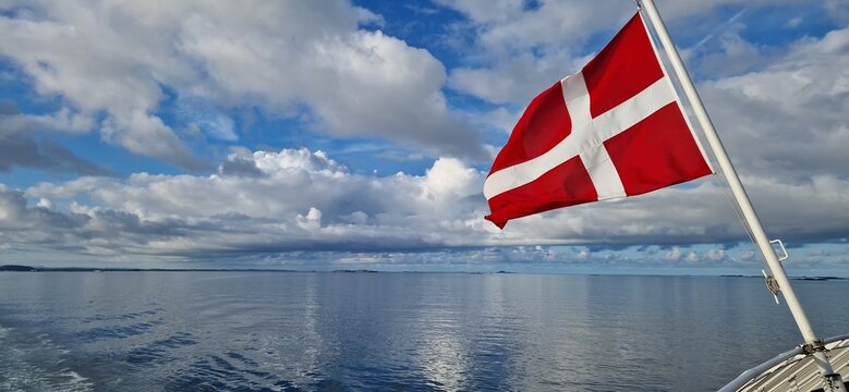 Danish national flag waving on a boat above calm waters under a dramatic cloudy sky. Travel, maritime transport and Scandinavian destination concept.