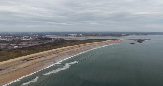 Aerial footage of the coastline near Redcar Middlesbrough showing beach sea waves and distant industrial area with port buildings across a wide landscape scene along the shore in the spring time