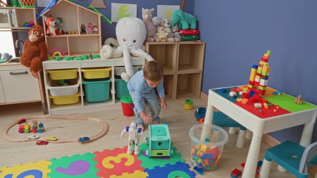 Little blond boy reaches to pick up a toy truck and manipulates a robot on a colorful foam number mat inside a playroom building; curiosity imagination.