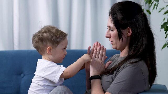Mother and toddler playing patty-cake on sofa. Mom claps hands with small child at home. Parent and baby boy bonding indoors. Mother teaches toddler clapping game. Family love and child development.