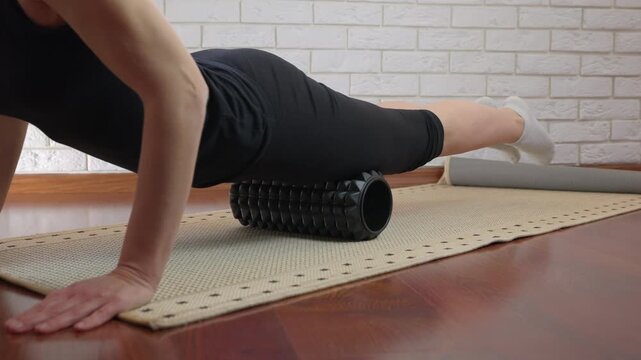 Woman performing myofascial release with foam roller. Athletic woman in black sportswear massaging her quadriceps with a foam roller on a mat, performing self-myofascial release for muscle recovery