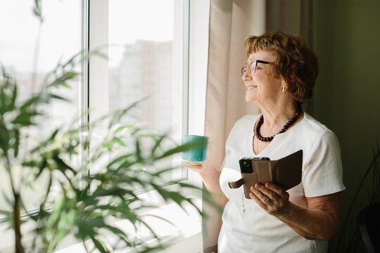 Senior woman smiling looking out window holding phone and mug