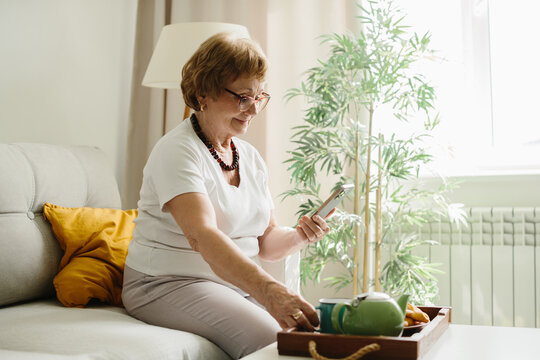 Senior woman relaxing on sofa using smartphone enjoying tea