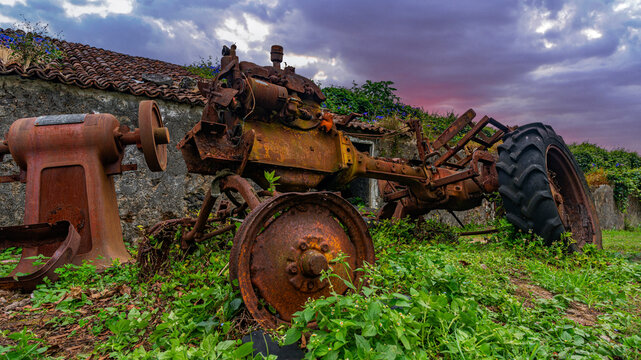 Rusty broken agricultural machinery abandoned outside ruined farmhouse at sunset