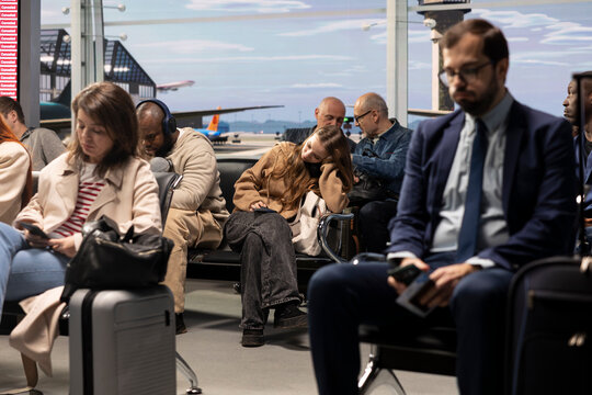 Bored men and women sitting on chairs inside busy airport lounge, waiting for plane to takeoff.