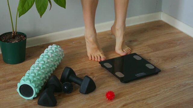 Woman pulls the toe of the foot, stretching her legs with bare feet when stepping on digital floor scales, weighing, checking at home near dumbbells, yoga roller after exercises, success dance.