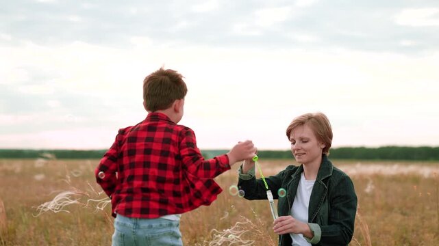 Woman blows bubbles while energetic boy catches in field in cloudy weather. Recreation and entertainment with bubbles for woman and boy in nature. Positive moment of woman and son playing with bubbles
