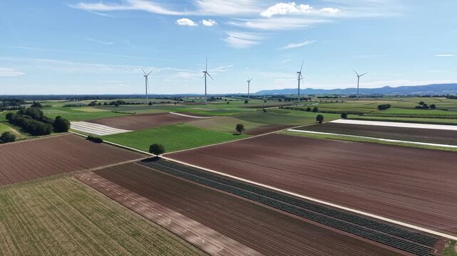 High-altitude drone video showing wind turbines rising above geometric agricultural fields. Modern renewable energy infrastructure integrated into productive farmland and open countryside