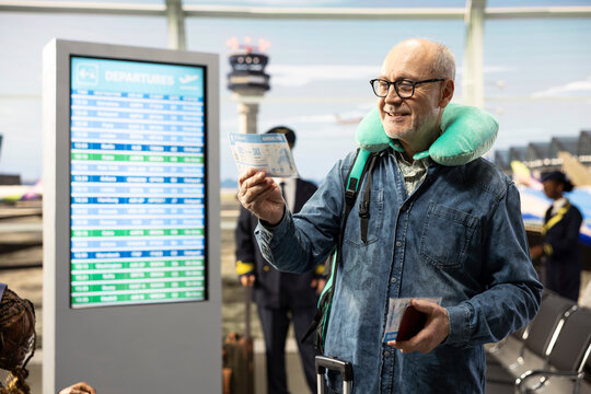 Happy older man reads boarding pass with travel pillow in airport lounge, smiling before flight departure.