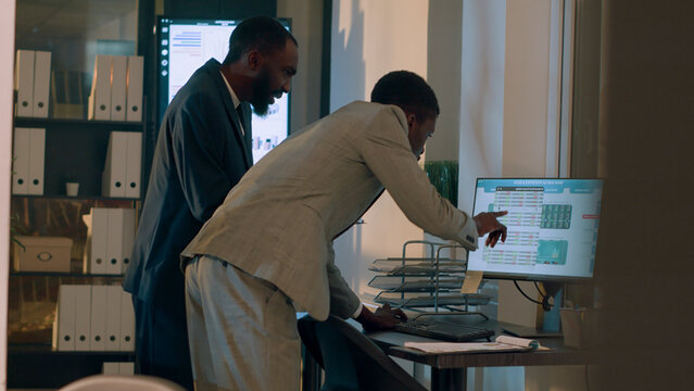 African American businessmen trying to troubleshoot reason for company revenue down from last year, checking numbers in office, employees looking over financial charts during night shift.
