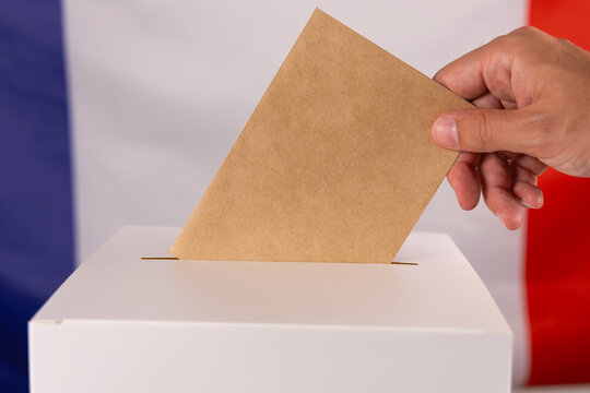 Gros plan sur la main d'une personne en train d'ins&eacute;rer un bulletin de vote dans une urne, pour les &eacute;lections. En arri&egrave;re plan, le drapeau de la France.
