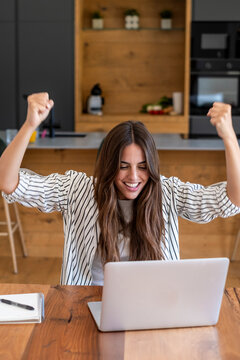 Happy young woman celebrating success at her laptop in a cozy kitchen workspace, raising her arms in triumph while working remotely, studying, or joining a video call