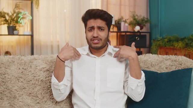 Indian man at home looking surprised, raising hands in a what why gesture, expressing confusion. Arabian guy sitting on sofa, reacting with disbelief, puzzled and questioning something unexpected