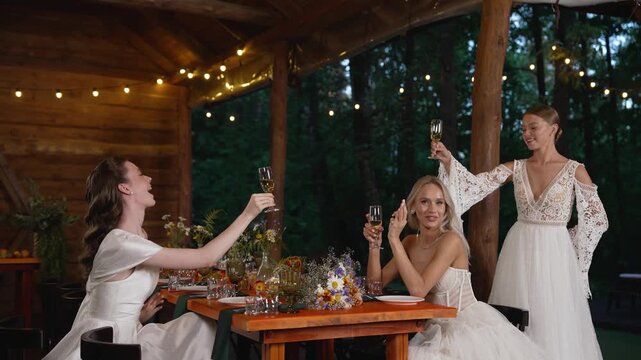 Three happy brides in white wedding dresses toasting with wine glasses at wooden table decorated with wildflowers under string lights in outdoor pavilion surrounded by forest
