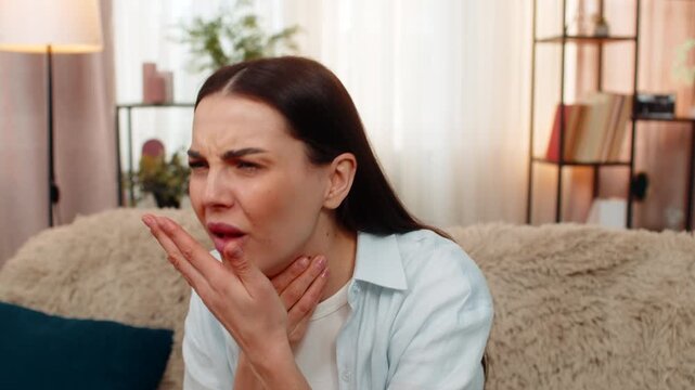 Young woman at home sitting on sofa holding throat feeling pain, coughing and looking sick. Caucasian sick girl experiencing flu symptoms, sore throat discomfort, unhealthy, showing illness signs