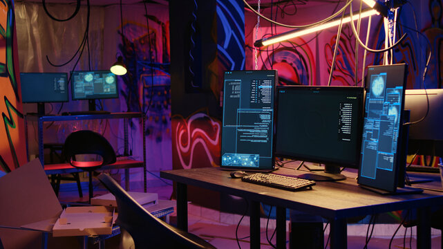 Computer screens showing malware development code in rundown underground hideout with graffiti painted on walls, PC system equipment used for cybercriminal activity in neon lit place.