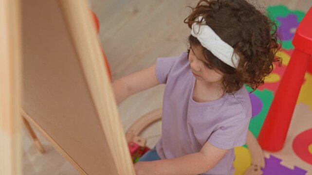 Little girl draws with purple chalk on easel, hand reaching across playmat inside building; playful curiosity.