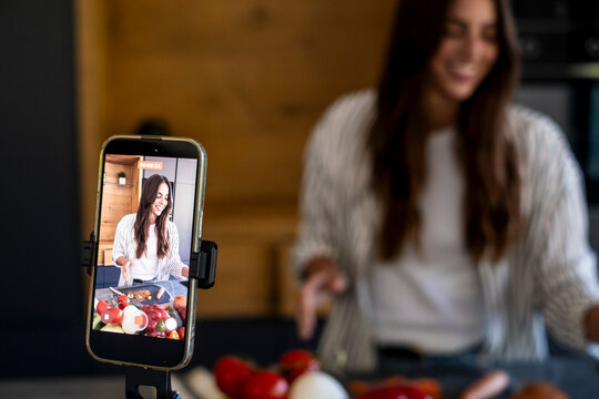 Young woman cooking and smiling while filming a food vlog on a smartphone in a modern kitchen, content creator demonstrating recipes and fresh ingredients