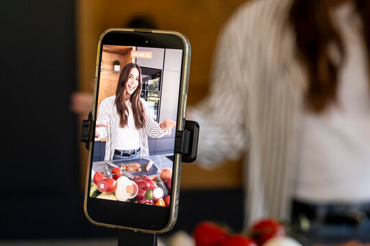 Young woman vlogger recording a cooking video on a smartphone in a modern kitchen with fresh vegetables on the counter, content creator demonstrating a recipe and engaging with viewers
