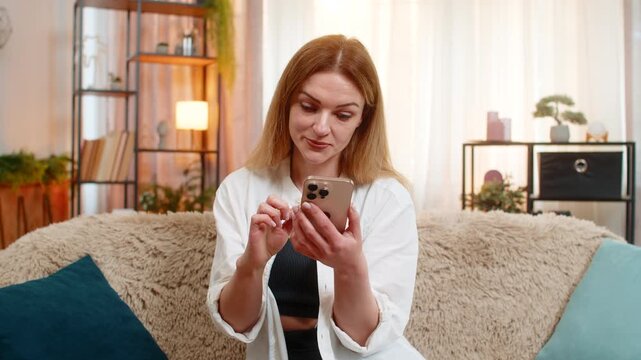 Middle-aged woman scrolls comments on smartphone at home, frowning and waving hand with frustration stress. Young adult sad girl shows nervous reaction to negative feedback, visibly annoyed and tense