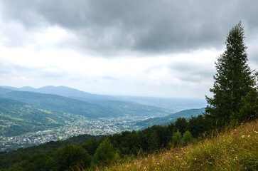Fototapeta premium Panoramic view of a Yaremche village nestled in a green mountain valley, surrounded by dense forests and rolling hills under a dramatic overcast sky. Carpathian Mountains, Ukraine