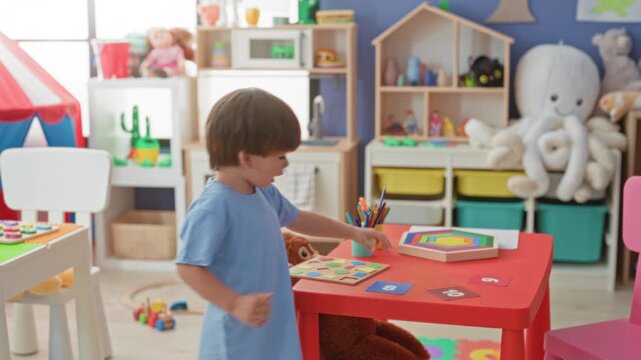 Boy plays with a wooden shape puzzle on a red table, reaching for crayons beside a stuffed monkey and colorful toy bins in a play area inside a building; childhood playfulness.