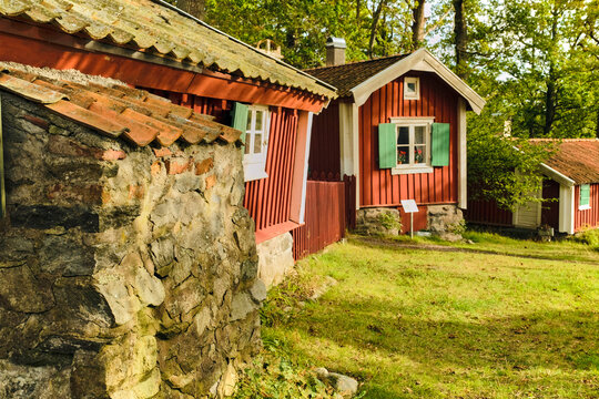 Karlskrona, Sweden - 13 September 2025: View of small historic cottages in Womoparken with red wooden walls, green shutters, and stone foundations surrounded by lush trees.