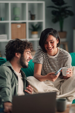 Cheerful couple spending time together on couch, woman holding phone and coffee mug, man laughing beside laptop