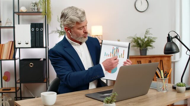 Middle-aged businessman at home office shows chart on laptop in remote call pointing at big drop. Freelancer guy at table proposes recovery plan gestures confident explaining steps for future growth.