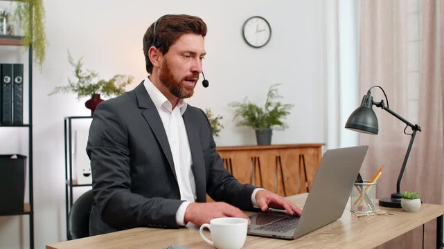 Middle-aged businessman at home office serves online hotline wears headset listens with patience and enters solution on laptop. Freelancer guy at table speaks warm tone confirms steps and logs notes.