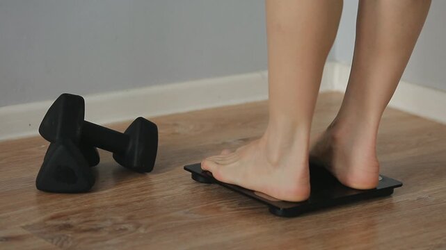 Woman bare feet stepping on black digital floor scales, she weighing, checking at home near , black sport dumbbells after exercises. Measuring weight, control, wellness, fat, body care. BMI concept