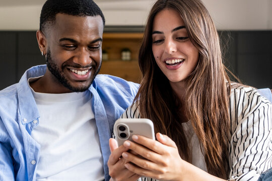 Young mixed-race couple sitting on a sofa smiling and looking at a smartphone together, sharing a moment of happiness, connection and casual home life