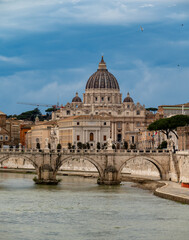 Fototapeta premium View on stone-arched bridge on Tiber river and St Peter's Basilica in Rome, cloudy sky, ochre and terracotta buildings, domes and umbrella pines rise on gently sloping Vatican hill