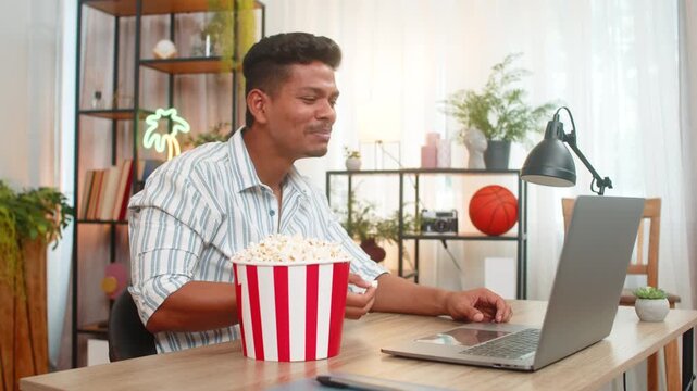 Indian man at home watches comedy film on laptop eats popcorn laughs and enjoys funny scene. Arabian handsome guy at desk smiles takes bite from backet looks at screen feels joy relax and happy mood