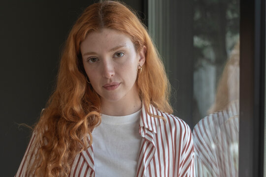 Young woman with red hair portrait in a striped shirt by a window with reflection, natural light and a thoughtful, calm expression