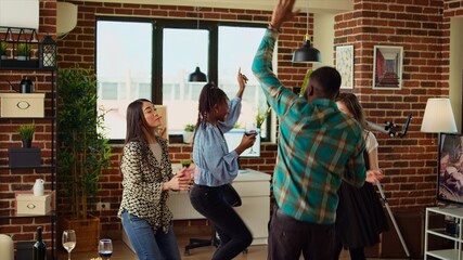 Multicultural youthful group of adults dancing at apartment party while drinking wine and champagne. Friends jumping around house, busting dance moves to energetic music in cozy living room