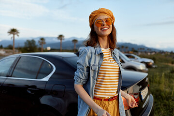 Fototapeta premium A cheerful woman in a striped shirt, denim jacket, orange beanie and sunglasses stands beside a parked car in a sunny outdoor setting. Lifestyle portrait of casual confidence.