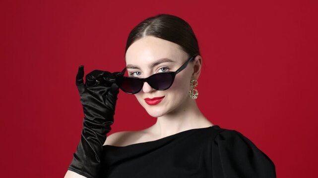Young woman wearing stylish sunglasses and black dress on red background