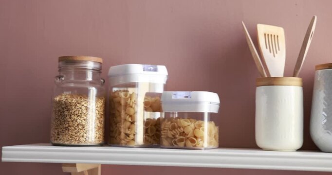 Kitchen utensils and food products on shelf near pink wall indoors, closeup. Camera moving left