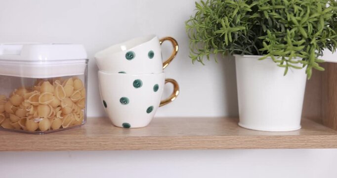 Food products, cups and plant on wooden shelf near white wall indoors, closeup. Camera moving right