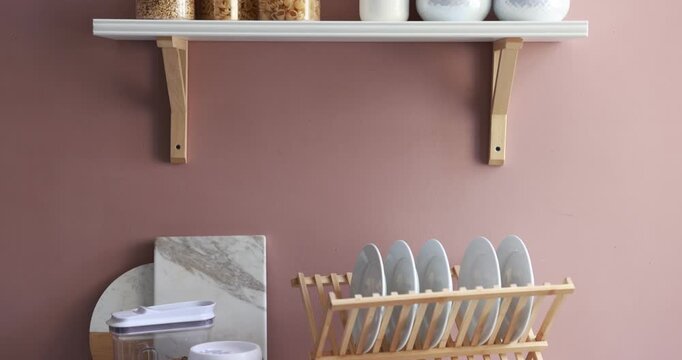 Shelf with food products and kitchen utensils near pink wall indoors, camera moving down
