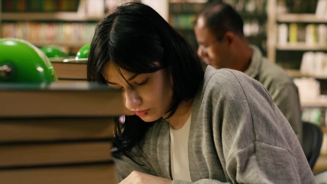 Focused woman student reading open book beside stack of books in library, study session with coursework under warm lighting.