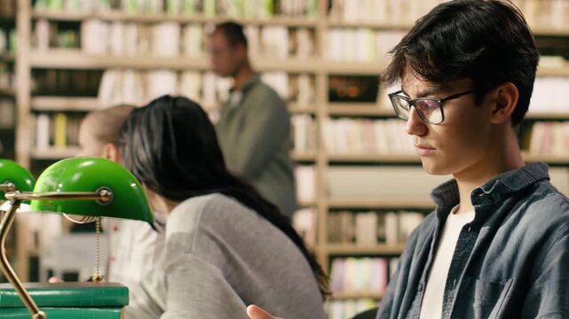 Portrait of young man reading open book at library study table for self study session, creating academic scene with stacked books near green lamps and bookshelves, Classmates study indoors.
