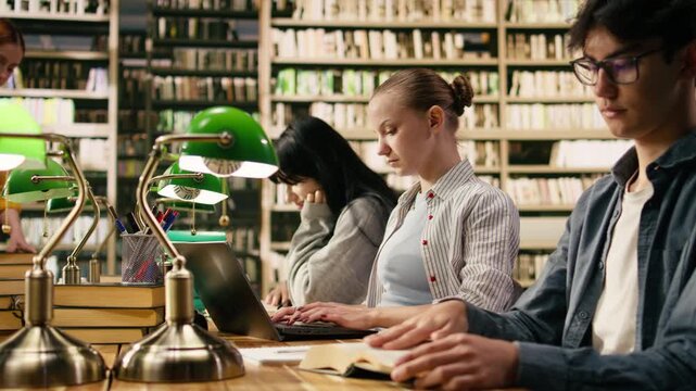 Student typing on laptop and taking notes from open book at library desk, reading coursework and study materials for academic research, Combining digital research with independent study.
