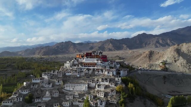 Thiksey Monastery rises from a hillside surrounded by green poplar trees and village buildings in the Indus Valley. Smooth drone shot reveals the white-washed Buddhist complex against barren&nbsp;mountains