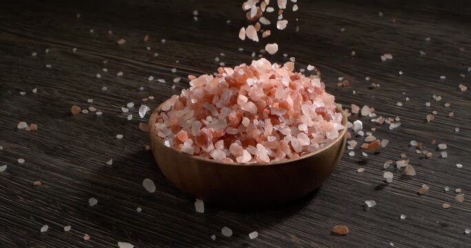 Slow motion close up of pink himalayan salt falling into a wooden bowl on a dark rustic wooden background for culinary concepts.